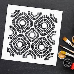 May include: A white stencil featuring a repeating geometric pattern of interlocking circles and squares in black. The stencil is on a black surface with paintbrushes and paint pots nearby, suggesting its use in art or craft projects.