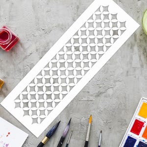 May include: A white stencil with a repeating geometric pattern of interlocking shapes. The stencil is on a gray surface, surrounded by art supplies including paintbrushes, watercolor paints, and small jars of paint.