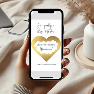 May include: A white smartphone with a gold heart on the screen. The text on the screen reads "J'ai quelque chose &agrave; te dire" and "VEUX-TU &Ecirc;TRE MON Marraine?" with the words "Marraine?" scratched off. The text at the bottom of the screen reads "GRATTEZ POUR D&Eacute;COUVRIR".