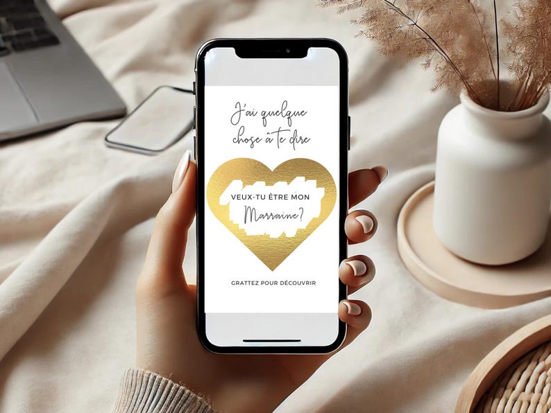 May include: A white smartphone with a gold heart on the screen. The text on the screen reads "J'ai quelque chose &agrave; te dire" and "VEUX-TU &Ecirc;TRE MON Marraine?" with the words "Marraine?" scratched off. The text at the bottom of the screen reads "GRATTEZ POUR D&Eacute;COUVRIR".