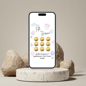 May include: A smartphone displays a gender reveal game with the text "Fille ou Gar&ccedil;on?" (Girl or Boy?). The screen features watercolor illustrations of a cloud, a onesie, a star, a pacifier, and hot air balloons. The game has golden scratch-off circles.