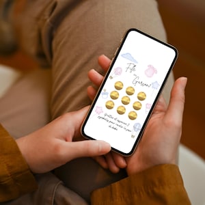 May include: A smartphone screen displays a baby gender reveal game. The screen features the text "Fille ou Gar&ccedil;on?" with illustrations of a cloud, a baby bottle, a star, a onesie, and a hot air balloon. There are 12 gold scratch-off circles.