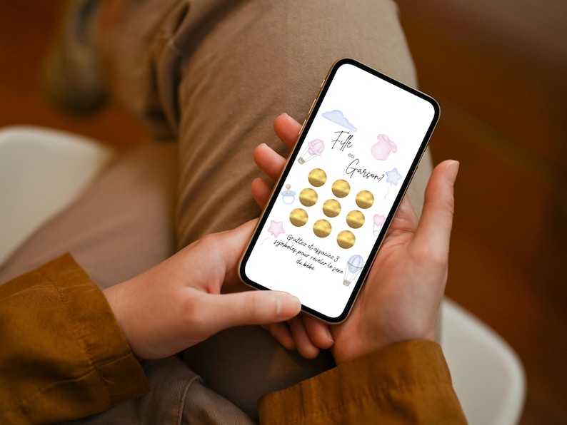 May include: A smartphone screen displays a baby gender reveal game. The screen features the text "Fille ou Gar&ccedil;on?" with illustrations of a cloud, a baby bottle, a star, a onesie, and a hot air balloon. There are 12 gold scratch-off circles.