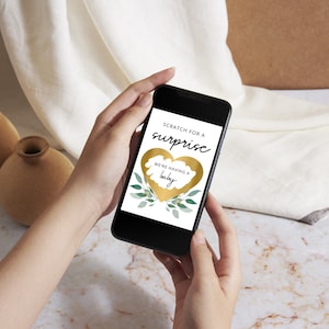 K&ouml;nnte beinhalten: Ein goldenes Herz mit einer Rubbelfl&auml;che auf dem Bildschirm eines Smartphones. Der Text auf dem Bildschirm lautet "Rubbeln Sie f&uuml;r eine &Uuml;berraschung" und "Wir bekommen ein Baby".