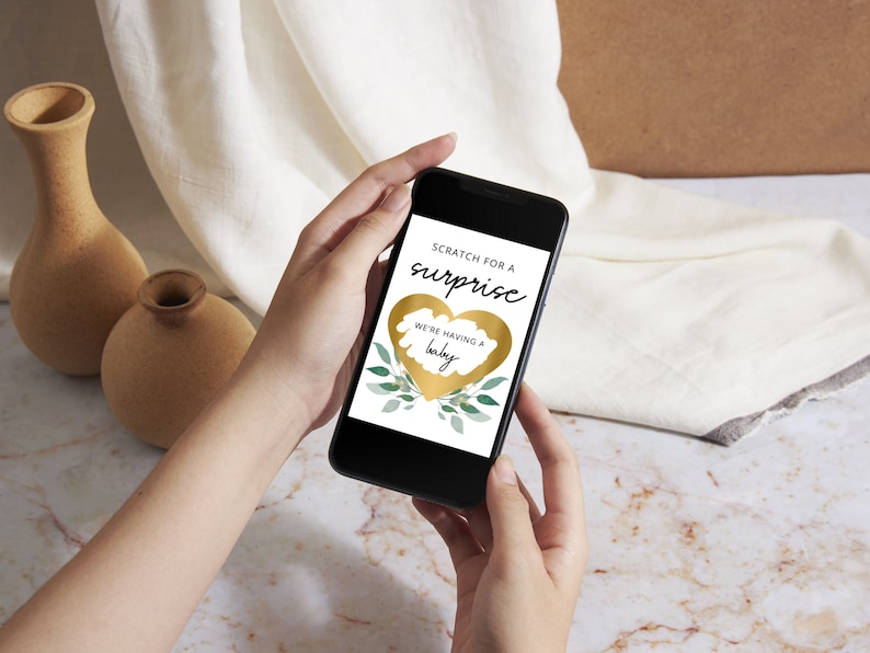 K&ouml;nnte beinhalten: Ein goldenes Herz mit einer Rubbelfl&auml;che auf dem Bildschirm eines Smartphones. Der Text auf dem Bildschirm lautet "Rubbeln Sie f&uuml;r eine &Uuml;berraschung" und "Wir bekommen ein Baby".