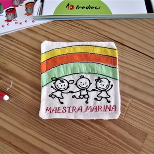 May include: A white fabric square with a rainbow design and three stick figures holding hands. The text "MAESTRA MARINA" is embroidered in red below the figures.
