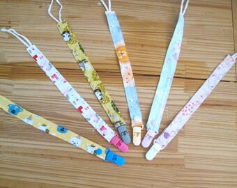May include: Six fabric pacifier clips featuring various patterns including koalas, jungle animals, and clouds. Each strap has a white cord loop at one end and a plastic clip in colors like blue, pink, gray, yellow, purple, and white at the other end.