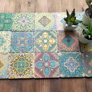 Könnte beinhalten: Eine Sammlung von vierzehn quadratischen Keramikfliesen, die in einem Raster angeordnet sind, jede mit einem einzigartigen, komplizierten, mehrfarbigen Blumen- und Geometriemuster in Grün-, Gelb-, Rosa- und Blautönen, begleitet von zwei kleinen Sukkulenten.