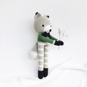 May include: A gray and white striped crocheted fox toy wearing a green shirt with a black bow tie. The fox is holding a small bouquet of white flowers.