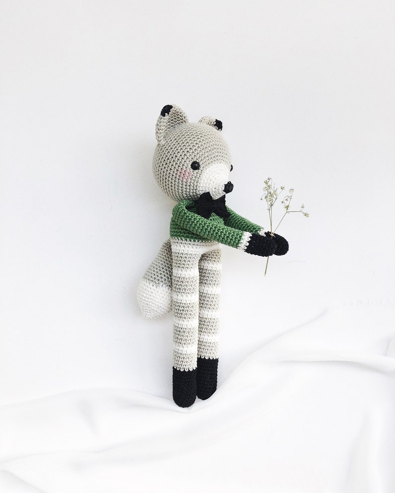 May include: A gray and white striped crocheted fox toy wearing a green shirt with a black bow tie. The fox is holding a small bouquet of white flowers.