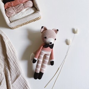 May include: A pink and white striped crocheted fox toy with black accents, wearing a black bow tie. The toy is sitting on a white surface with two dried grass stems.