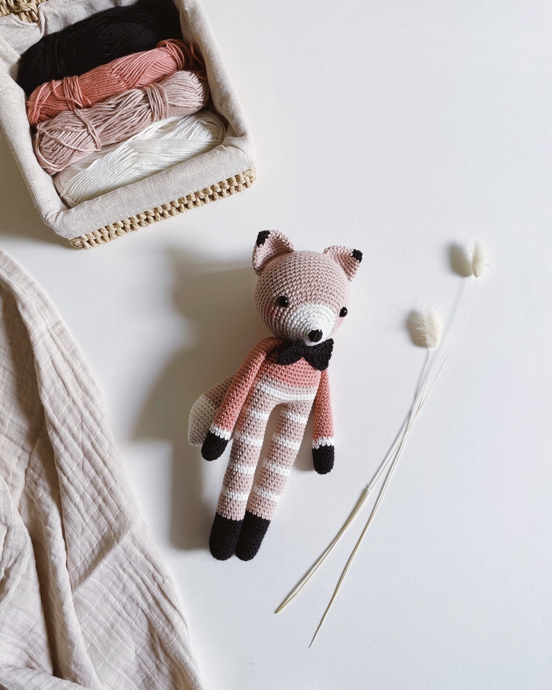May include: A pink and white striped crocheted fox toy with black accents, wearing a black bow tie. The toy is sitting on a white surface with two dried grass stems.