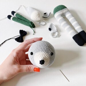 May include: A crocheted animal toy in progress. The head is complete with black eyes and a black nose. The legs are striped grey and white with green at the top. The ears are grey and white with black trim. A black bow tie and a needle are also visible.