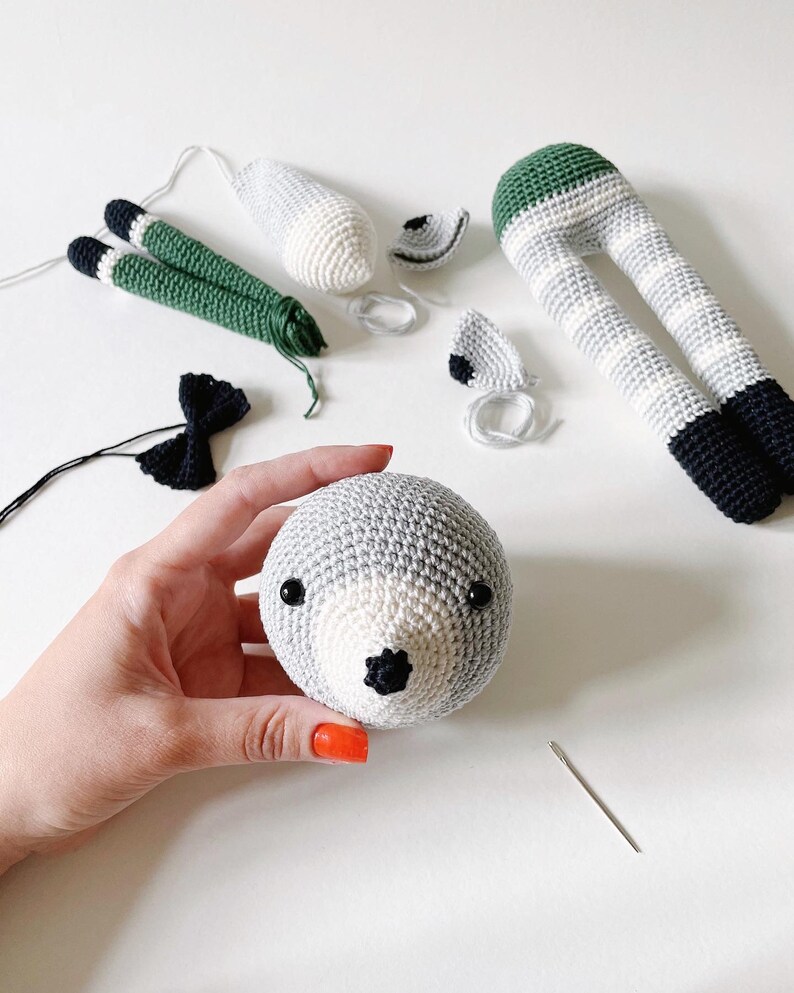 May include: A crocheted animal toy in progress. The head is complete with black eyes and a black nose. The legs are striped grey and white with green at the top. The ears are grey and white with black trim. A black bow tie and a needle are also visible.