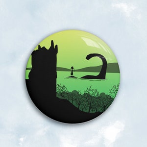 Peut inclure: Un bouton rond avec une illustration fantaisiste d'une personne debout dans l'eau face à une grande créature, peut-être le monstre du Loch Ness. Une silhouette de château est à gauche, avec un fond dégradé vert et jaune. Le bouton a une finition brillante.