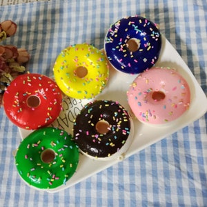 May include: Six colorful donuts with sprinkles on a white plate. The donuts are red, yellow, blue, pink, green, and brown.