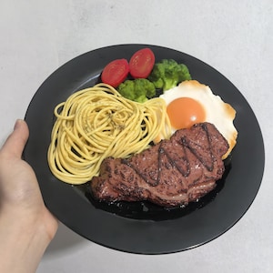 Puede incluir: Un plato negro con un filete, espaguetis, un huevo frito y guarnición de brócoli y tomate.