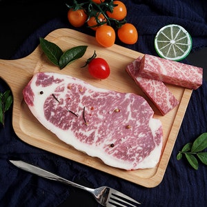 May include: A raw, marbled beef steak on a wooden cutting board with a few sprigs of green herbs, a lime wedge, and cherry tomatoes.