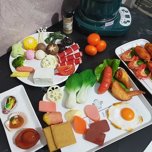 Puede incluir: Un plato blanco con una variedad de alimentos falsos coloridos, incluyendo verduras, carne y mariscos. La comida está dispuesta de forma visualmente atractiva, y el plato está rodeado de otros platos de comida.