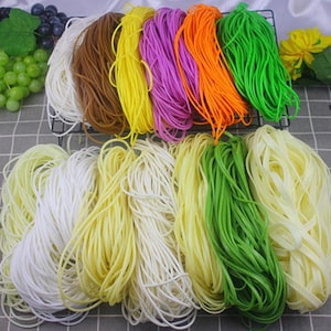 Puede incluir: Surtido de cordones elásticos de colores en varios colores, incluyendo blanco, amarillo, verde, naranja, morado, marrón y azul claro. Los cordones están dispuestos en filas y están listos para ser utilizados en proyectos de manualidades.