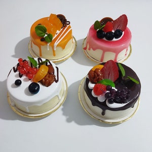 May include: Four miniature cakes with various toppings are displayed on a white surface. Each cake has a different topping, including orange slices, strawberries, blueberries, and chocolate. The cakes are on small, gold-colored paper plates.