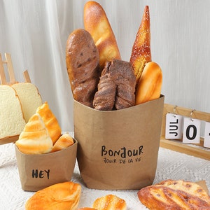 May include: A brown paper bag filled with various types of bread, including baguettes, croissants, and rolls. The bag has the text "Bonjour Jour De La Vie" printed on it. There are other bread items on a table in front of the bag.