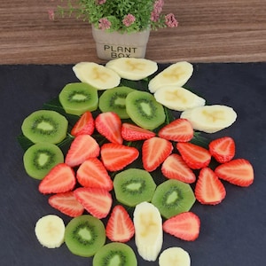 Puede incluir: Una colorida disposición de fruta en rodajas, que incluye kiwi verde, fresas rojas y rodajas de plátano amarillas. La fruta está dispuesta sobre una superficie oscura, con una pequeña planta en maceta en el fondo. La maceta tiene el texto "PLANT BOX".