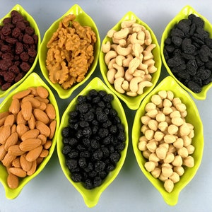 May include: Seven small green bowls filled with different types of nuts and dried fruit. The bowls are arranged in a row on a light blue surface. The nuts and dried fruit include almonds, cashews, walnuts, raisins, and dried cranberries.