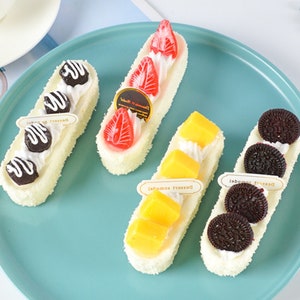 Puede incluir: Cuatro modelos de postre falsos en un plato turquesa. Los postres son todos largos y rectangulares, con glaseado blanco y diferentes coberturas. Uno tiene chispas de chocolate y un chorrito de chocolate, otro tiene fresas, otro tiene mango y otro tiene galletas Oreo.