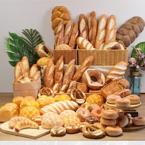 Puede incluir: Una variedad de productos horneados, que incluyen baguettes, panes, pretzels y donas, están dispuestos sobre una superficie de madera. Algunos artículos se exhiben en cestas de mimbre y en un soporte escalonado. El surtido presenta varias formas y colores.