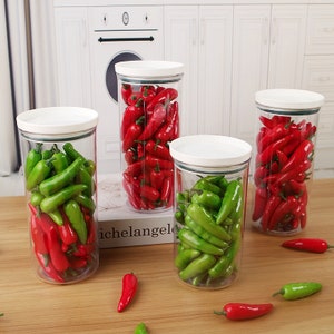 Puede incluir: Cuatro tarros de vidrio transparente con tapas blancas llenos de chiles rojos y verdes. Los tarros están sobre una superficie de madera.