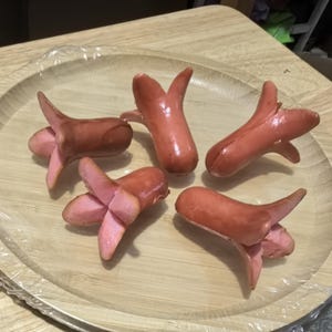 Puede incluir: Cinco salchichas rosas cocidas dispuestas en un plato de madera. Cada salchicha ha sido cortada para crear una forma de flor, con los extremos divididos en pétalos. El plato está cubierto con film transparente.