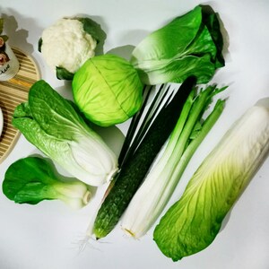 May include: Assortment of artificial vegetables, including a green cabbage, a white cauliflower, a green cucumber, a bunch of green onions, and three different types of green lettuce.