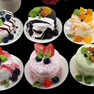 Pode incluir: Seis pilhas de panquecas em miniatura decoradas com diferentes coberturas, incluindo frutas, chocolate e granulado. As panquecas são exibidas em pratos brancos.