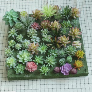 Puede incluir: Una base cuadrada verde cubierta con un surtido de plantas suculentas artificiales en varios tonos de verde, rosa, amarillo y morado. Las plantas presentan diversas formas de hojas, incluyendo rosetas, follaje puntiagudo y texturas redondeadas.