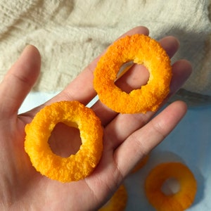 May include: Close-up of a hand holding four orange and yellow cheese puffs shaped like rings. The puffs are a popular snack food.