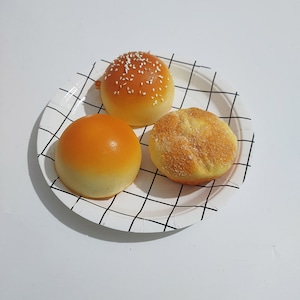 May include: Three round bread rolls on a white plate with a black grid pattern. The rolls are all different shapes and sizes. One roll has sesame seeds on top.
