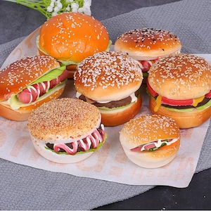 Peut inclure: Six différents types de pains à hamburger avec diverses garnitures, notamment de la laitue, de la tomate et du fromage. Les pains sont tous blancs et ont des graines de sésame sur le dessus.