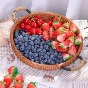 May include: A round woven basket filled with an assortment of artificial fruit. The basket contains cherries, strawberries, and blueberries. The basket has rope handles and sits on a printed paper surface.
