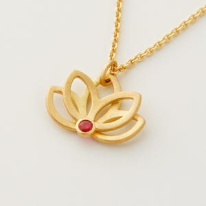 Peut inclure: Un collier pendentif doré avec un motif de fleur de lotus et une petite pierre précieuse rouge ronde au centre. La chaîne est fine. Le pendentif lotus mesure environ 2,5 cm de diamètre, sur fond blanc.