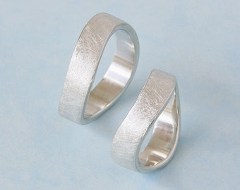 May include: Two silver wedding bands with a brushed finish. The bands are both slightly curved and have a unique, modern design.