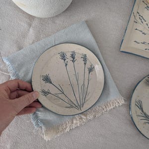 Könnte beinhalten: Ein runder Keramikteller mit cremefarbener Basis, gesprenkelter Textur und einer dunkelblauen botanischen Illustration von Lavendelzweigen. Der Teller hat einen dünnen dunkelblauen Rand und liegt auf einer hellblauen Stoffserviette mit Fransen.