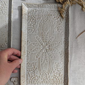 Könnte beinhalten: Ein rechteckiges Keramiktablett mit einem komplizierten, erhabenen, spitzenartigen Blumenmuster in Off-White- und Beigetönen. Die Oberfläche hat ein strukturiertes, gesprenkeltes Finish mit einem leicht unebenen Rand.