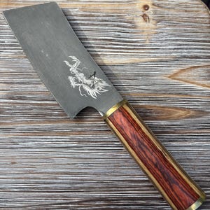 Könnte beinhalten: Ein rechteckiges Hackmesser mit einer silbernen Klinge und einem Holzgriff. Die Klinge zeigt ein weißes Drachendesign. Der Griff ist eine Kombination aus rotbraunem Holz und Messingakzenten. Das Messer ist für den Küchengebrauch konzipiert.