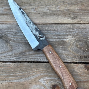 May include: A handcrafted chef's knife with a rustic, textured blade and a wooden handle. The blade is silver and the handle is a warm brown, secured with three silver rivets. The knife rests on a wooden surface.