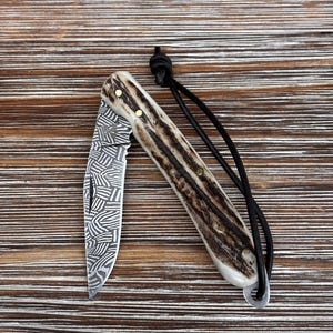 Può includere: Un coltello pieghevole con una lama a fantasia e un manico in materiale marrone e crema. Il coltello ha un cordino nero attaccato. La lama è aperta, rivelando un design dettagliato.