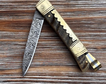 May include: A folding pocket knife with a Damascus steel blade featuring a wavy pattern. The handle is constructed from dark, textured horn material with brass bolsters and a decorative scalloped brass spine. A metal clip is attached to the end.