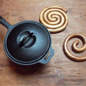 May include: A black cast iron pot with a lid and a wooden spiral trivet. The pot has a handle on the side and a loop handle on the lid.