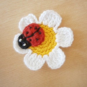 Könnte beinhalten: Eine gehäkelte Blume mit weißen Blütenblättern und einer gelben Mitte. Ein roter Marienkäfer mit schwarzen Punkten befindet sich in der Mitte der Blume.
