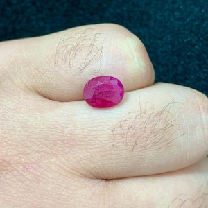 Puede incluir: Una vibrante piedra preciosa de rubí de corte ovalado con un tono rojo rosado intenso y una superficie facetada que refleja la luz. La piedra se muestra sobre un fondo oscuro, destacando su calidad translúcida y su forma ovalada pulida.